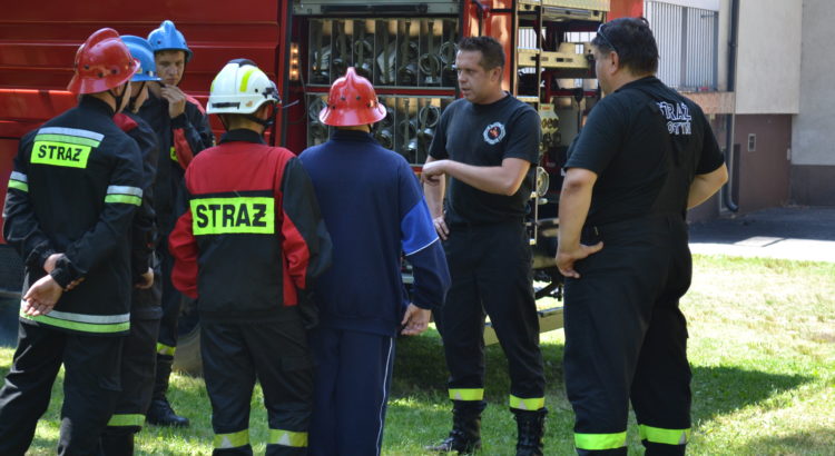 Dwóch strażaków z OSP Gostyń prowadzi szkolenie, wokół nich stoi młodzież, a za nimi widać samochód pożarniczy.